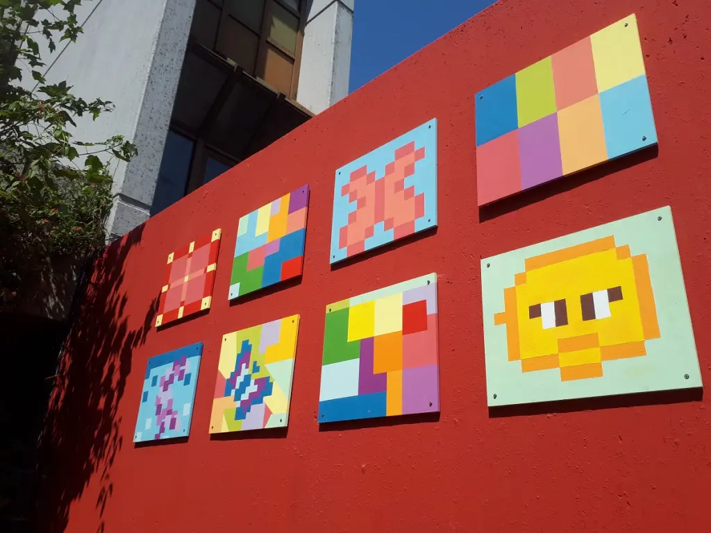 Auf dm Bild sieht man eine Außenaufnahme vor eine rot gestrichenen Wand. An der Wand befinden sich acht bunte, mosaikförmig angebrachte Bilder.