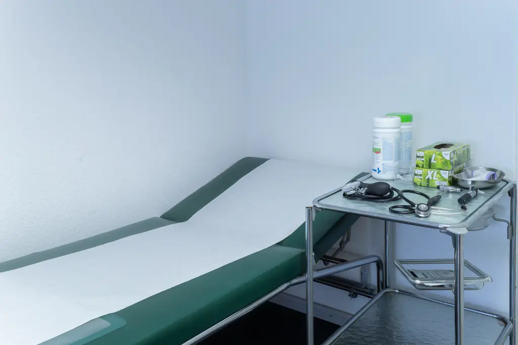 Foto: Eine Krankenhausliege mit einer Schutzfolie aus Pappe und einem Seitentisch mit Desinfektionsmittel und Latex-Handschuhen.