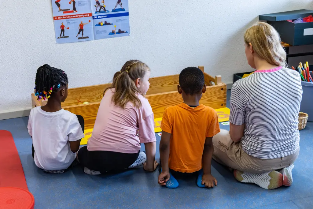 Drei Kinder sitzen neben einer erwachsenen Person, die Kinder sind von hinten zu sehen. Gemeinsam lösen sie eine Aufgabe.