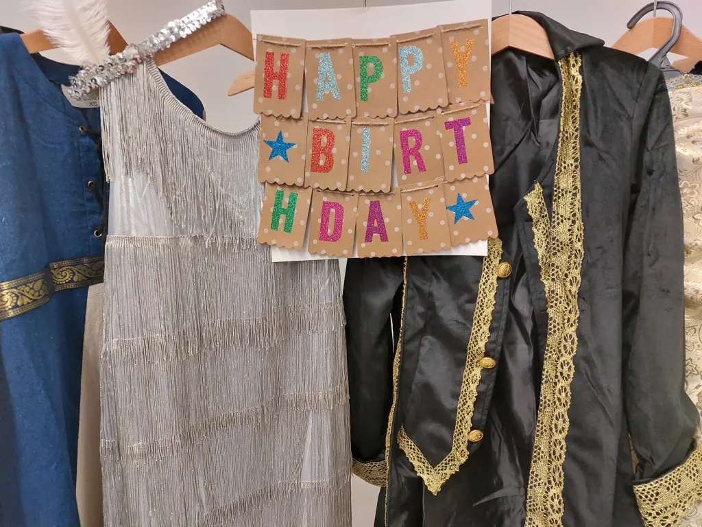 Foto: Ein Schild mit der Aufschrift "Happy Birthday" hängt neben historischen Kleidungsstücken auf einer Stange.