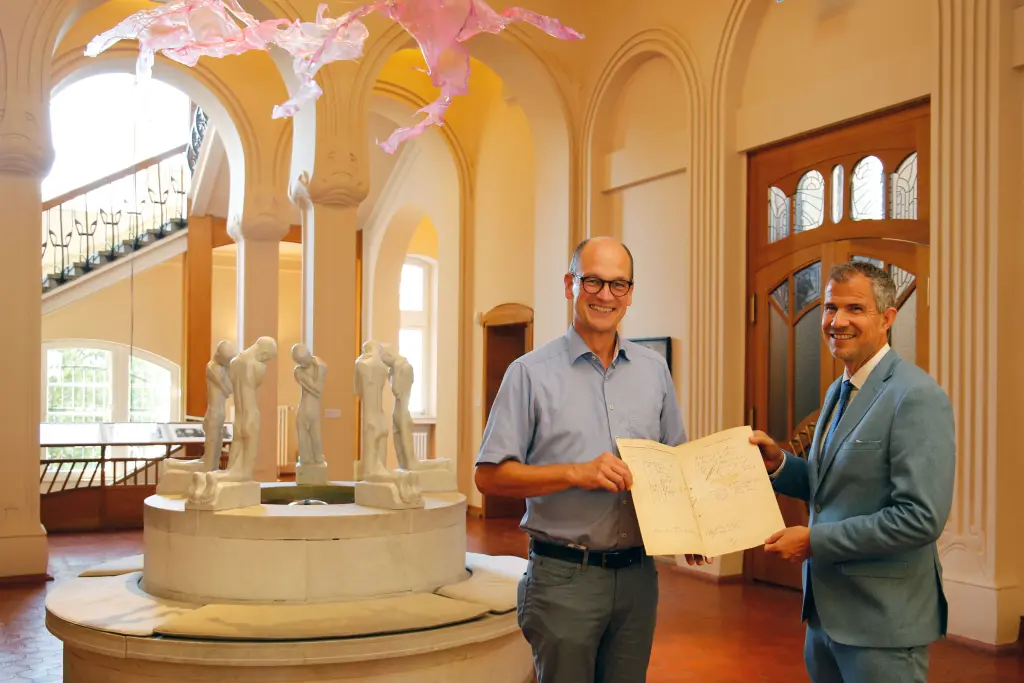 Auf dem Bild sind zwei Männer zu sehen. Sie stehen sich einander zugewandt. Gemeinsam halten sie in ihrer Mitte ein Schriftstück. Im Hintergrund des Bildes ist ein Museumssaal im Jugendstil zu sehen. In der linken Bildhälfte ist ein Brunnen aus Marmor.