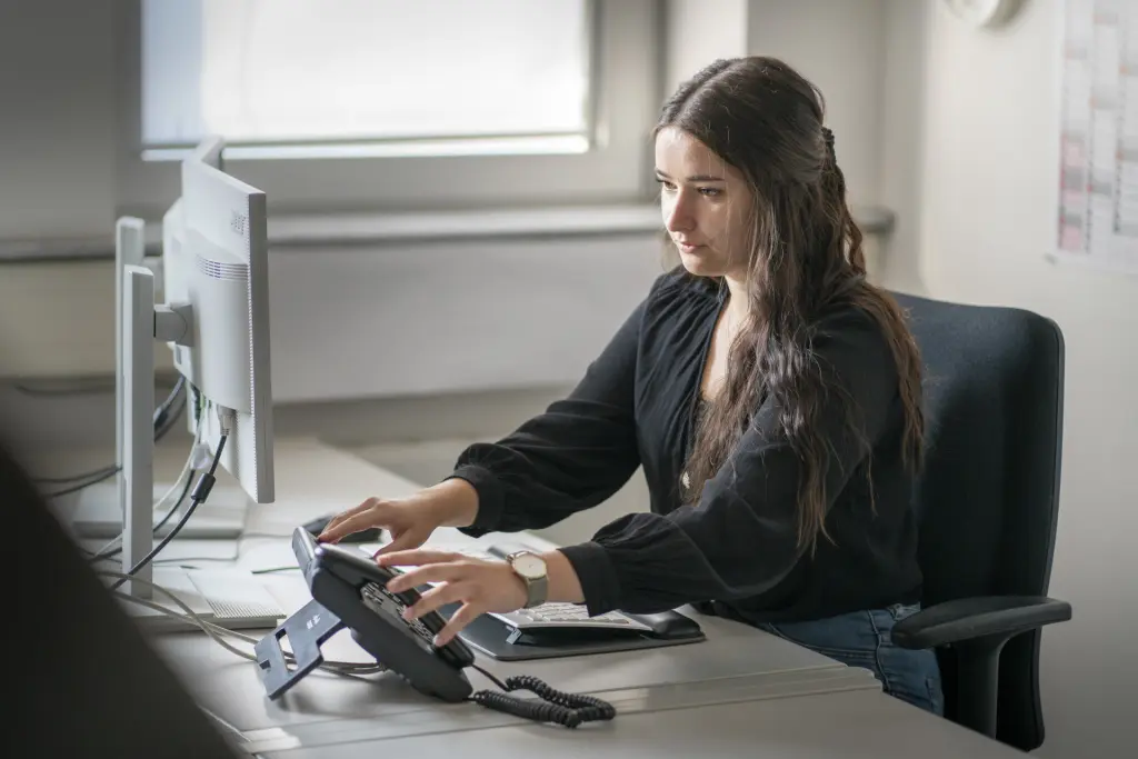 Eine Kollegin mit dem Abschluss Bachelor of Laws in Allgemeiner Verwaltung sitzt an ihrem Arbeitsplatz, hält die Hand am Telefonhörer, um das Gespräch anzunehmen, und blickt dabei auf ihren Bildschirm. Die Szene zeigt eine typische Arbeitssituation im Verwaltungsalltag.