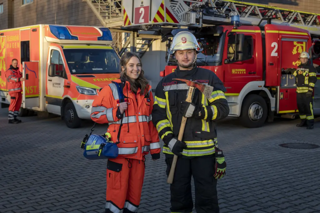 Eine angehende Notfallsanitäterin und ein Brandmeisteranwärter der Feuerwehr Hagen stehen vor einem Einsatzfahrzeug und einem Rettungswagen. Beide tragen ihre jeweilige Schutzkleidung und Ausrüstung. Im Hintergrund sind weitere Einsatzkräfte und Einsatzfahrzeuge der Feuerwehr Hagen zu sehen. Die Szene vermittelt Teamarbeit, Einsatzbereitschaft und die enge Zusammenarbeit zwischen Rettungsdienst und Feuerwehr.