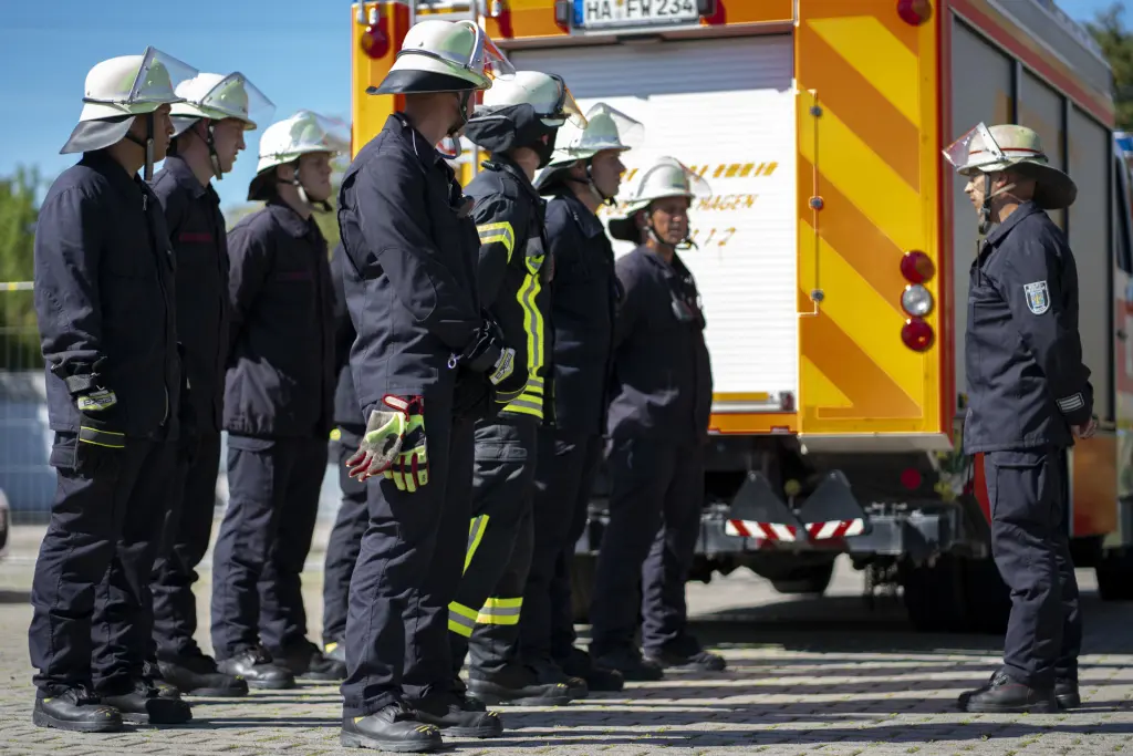 Eine Führungskraft der Feuerwehr steht vor einer kleinen Gruppe von Feuerwehrleuten und hält eine Besprechung. Im Hintergrund ist ein Feuerwehrfahrzeug des Typs HLF (Hilfeleistungslöschgruppenfahrzeug) zu sehen. Die Szene vermittelt Teamarbeit, Führungskompetenz und die wichtige Rolle der Einsatzplanung im Feuerwehrdienst.