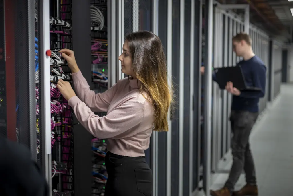 Eine Frau mit dem Abschluss Bachelor of Science in Verwaltungsinformatik – E-Government arbeitet mit beiden Händen an verschiedenen Kabeln vor einem Serverschrank. Im Hintergrund ist ein großes Rechenzentrum zu erkennen, in dem ein Kollege mit einem Laptop arbeitet. Die Szene zeigt praktische IT-Arbeit und die Infrastruktur im Bereich E-Government.