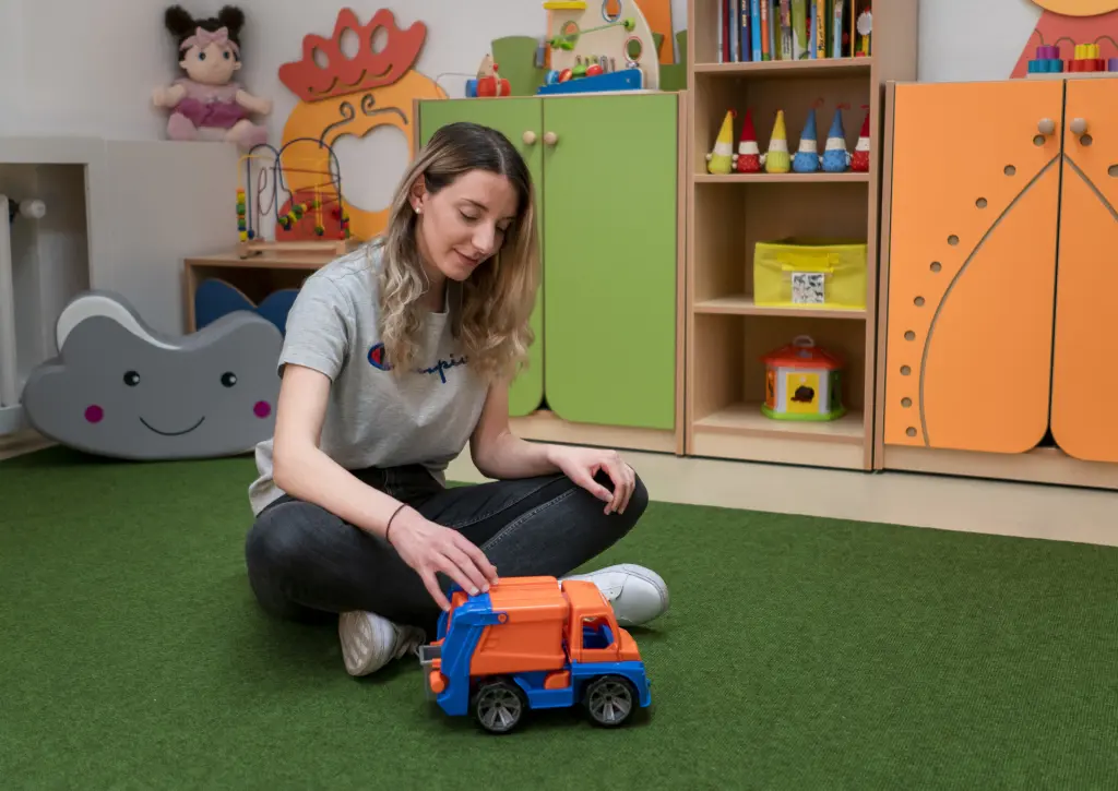 Eine angehende staatlich anerkannte Erzieherin (PiA) sitzt auf einem Spielteppich in einer Kindertagesstätte und spielt mit einem Modellauto. Die Aufnahme zeigt die engagierte pädagogische Arbeit im frühkindlichen Bildungsbereich.