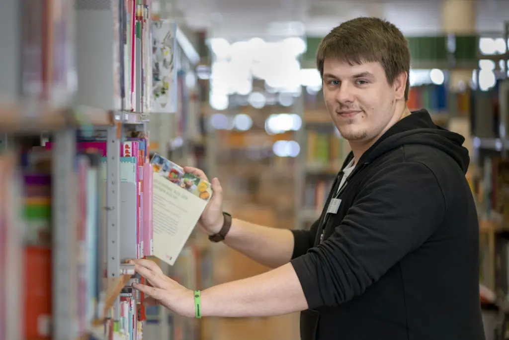 Ein Fachangestellter für Medien- und Informationsdienste, Fachrichtung Bibliothek, zieht ein Buch aus dem Regal und schaut dabei freundlich in die Kamera. Die Aufnahme entstand in der städtischen Bücherei und vermittelt den freundlichen Service und die Arbeit mit Medienbeständen.