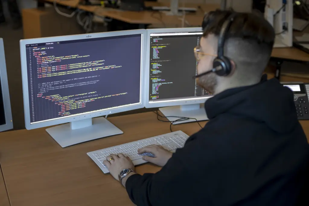 Ein Fachinformatiker für Anwendungsentwicklung sitzt mit einem Kopfhörer vor einem Computerbildschirm und betrachtet konzentriert Programmiercode. Die Szene zeigt die Arbeit an Softwareentwicklung und Coding in einem modernen Arbeitsumfeld.