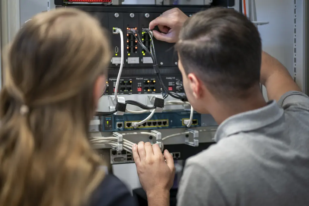 Eine männliche und eine weibliche Ausbildungskraft zur/zum Fachinformatiker*in der Fachrichtung Systemintegration stecken gemeinsam Kabel in ein Patchfeld. Die Aufnahme zeigt die praktische Arbeit im Bereich Netzwerktechnik und Systemadministration aus der Perspektive von hinten.