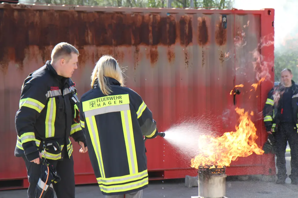 Foto: Eine junge Frau löscht einen kleinen Brand mit einem Feuerlöscher, daneben steht ein Feuerwehrmann.