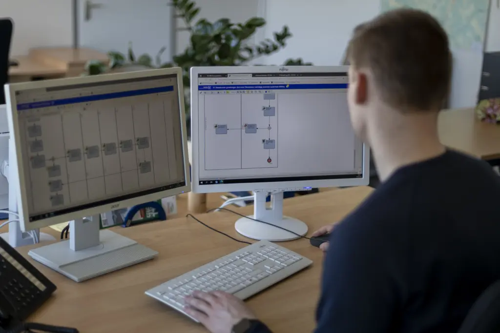 Ein Mann ist von hinten zu sehen und blickt auf zwei Bildschirme, auf denen verschiedene Verwaltungsprozesse visualisiert sind. Die Szene zeigt die Arbeit mit IT-Systemen zur Steuerung und Optimierung von Verwaltungsabl&auml;ufen.