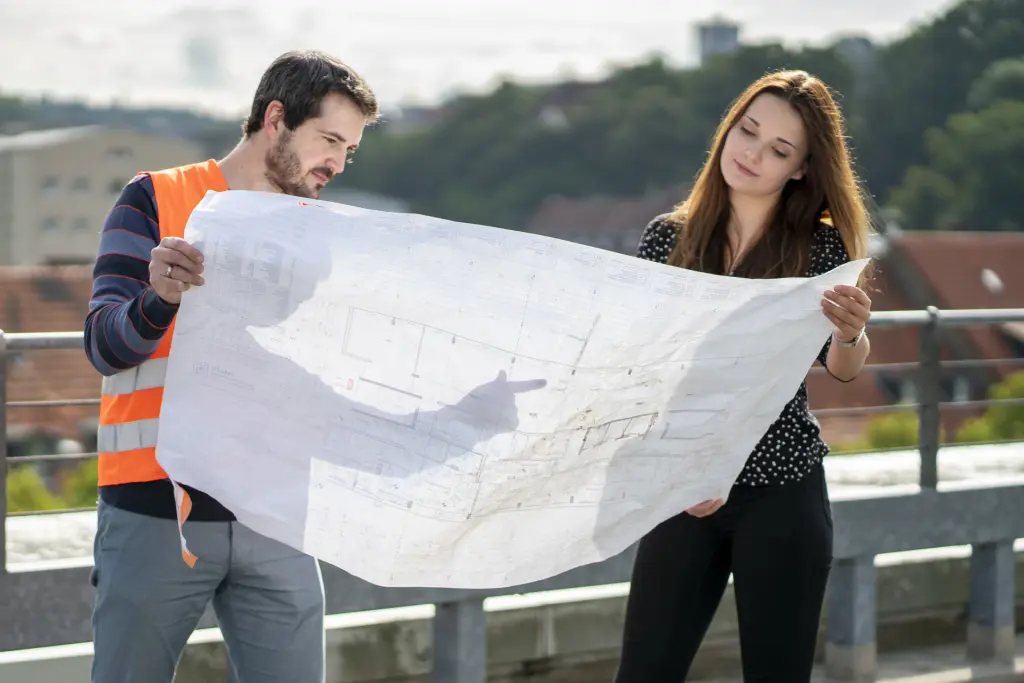 Ein m&auml;nnlicher Bauoberinspektoranw&auml;rter in auff&auml;lliger Warnweste steht konzentriert auf einer Baustelle und betrachtet aufmerksam einen Bauplan. Die Umgebung vermittelt einen praktischen Einblick in den Baualltag, w&auml;hrend der Kollege durch seinen fokussierten Blick Engagement und Fachwissen ausstrahlt.