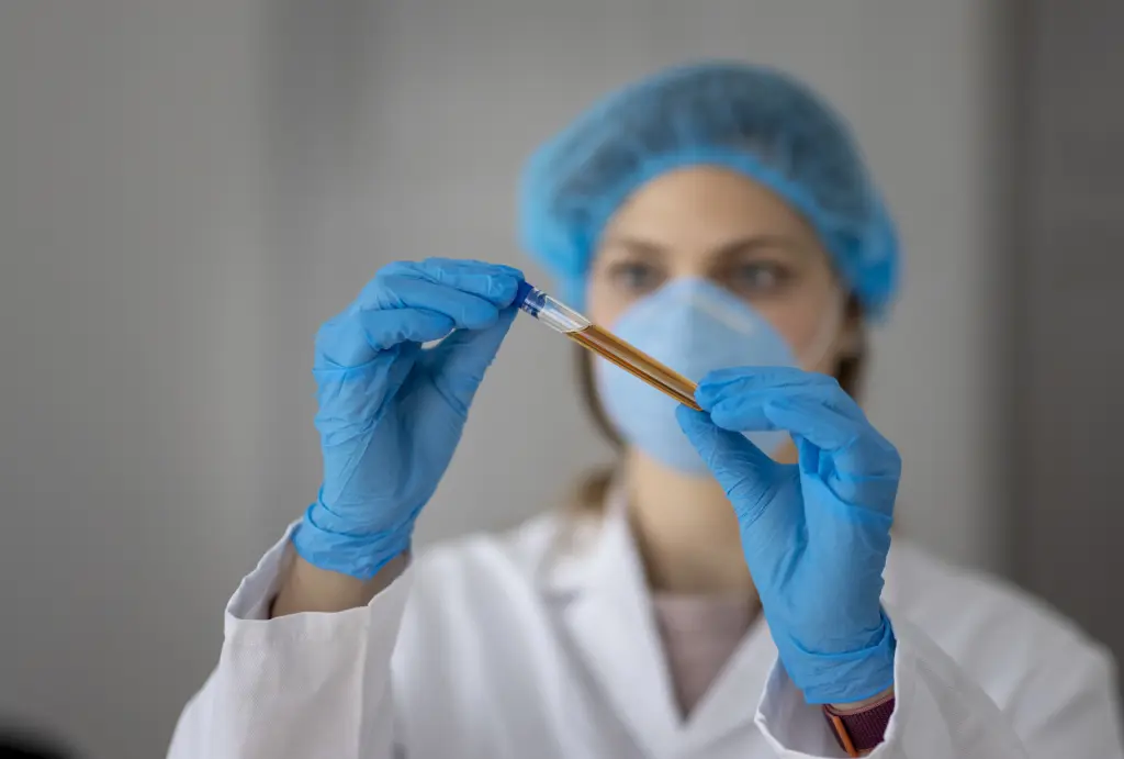 Eine Kollegin in Schutzkleidung untersucht aufmerksam eine Probe in einem Reagenzglas. Die Szene unterstreicht sorgf&auml;ltiges Arbeiten und den verantwortungsvollen Umgang mit Umweltproben, die f&uuml;r die Analyse und den Schutz unserer Umwelt von gro&szlig;er Bedeutung sind.