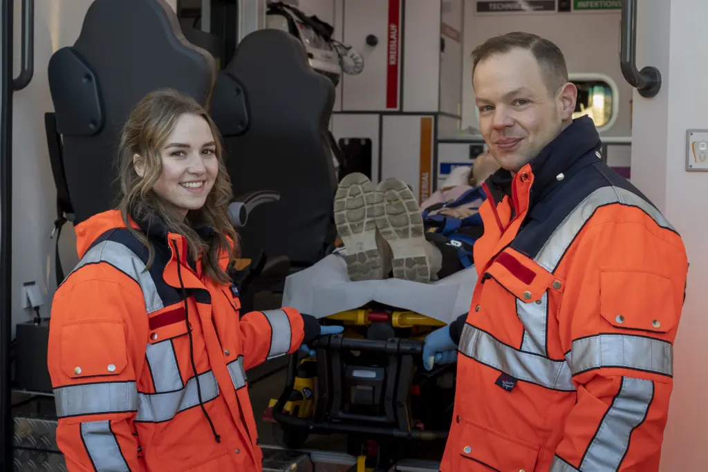 Zwei angehende Notfallsanitäter*innen, eine weiblich und eine männlich, verladen gemeinsam eine Patientin auf einer Trage in einen Rettungswagen (RTW). Die Szene zeigt den Einsatz und die Zusammenarbeit im Rettungsdienst bei der Patientenversorgung.