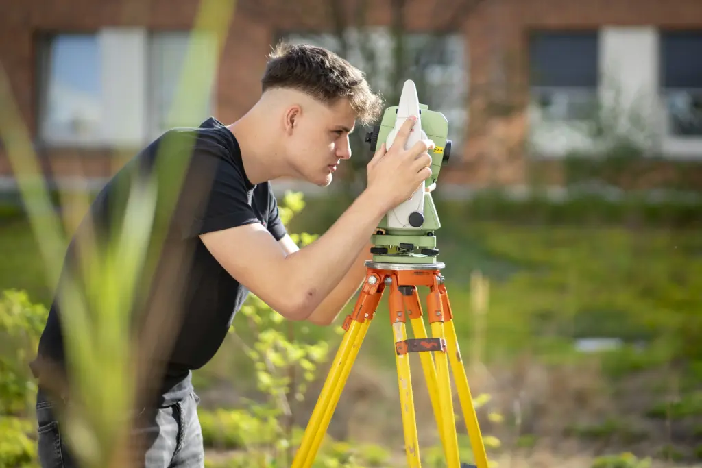 Ein männlicher Kollege blickt konzentriert durch sein Vermessungsgerät und visiert einen Messpunkt durch die Optik an. Die Aufnahme zeigt die Arbeit im Freien und verdeutlicht präzises, technisches Vorgehen bei der Geländeaufnahme und Vermessung.