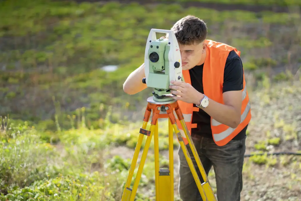 Ein Vermessungstechniker mit Warnweste führt im Außengelände eine Vermessung mit moderner Lasertechnik durch und blickt dabei durch seine Visieroptik. Die Szene zeigt präzise Vermessungsarbeiten im Außeneinsatz.