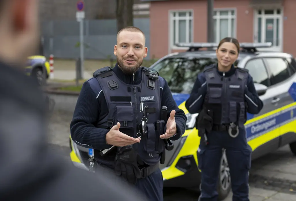 Ein Kollege aus dem städtischen Ordnungsdienst spricht mit einem Bürger und nimmt dabei eine freundliche, offene Haltung mit geöffneten Armen ein. Im Hintergrund sind seine Kollegin sowie ein Dienstfahrzeug des städtischen Ordnungsdienstes zu sehen. Die Szene zeigt den respektvollen und serviceorientierten Umgang im öffentlichen Dienst.