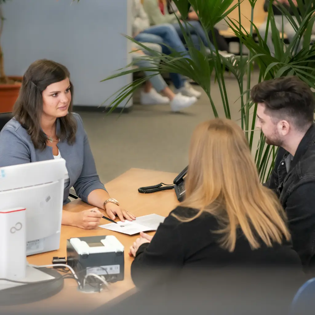 Eine Verwaltungswirtin berät ein Paar im Bürgeramt zu einem Antrag. Die Szene zeigt eine professionelle und freundliche Beratungssituation im öffentlichen Dienst.