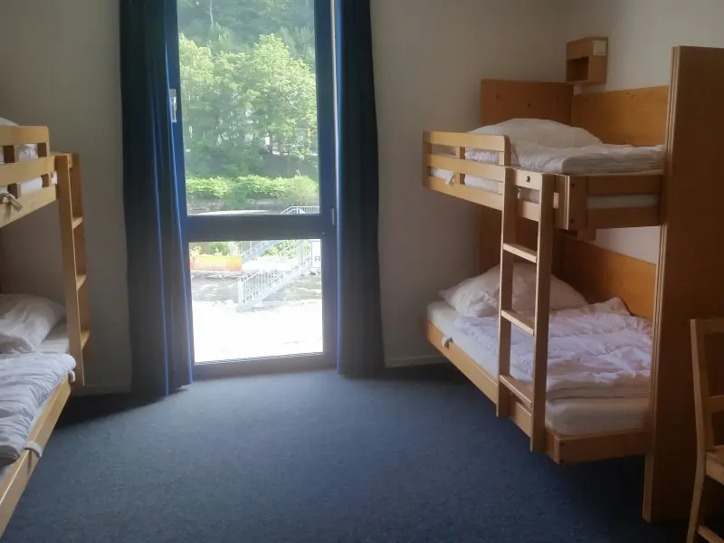 Foto: Ein zimmer mit einem Etagenbett, einem bodentiefen Fenster, einem Tisch mit Stuhl und Teppichboden.
