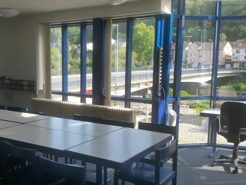 Foto: Ein Aufenthaltsraum mit bodentiefen Fenstern und einer langen Tischreihe mit Stühlen.