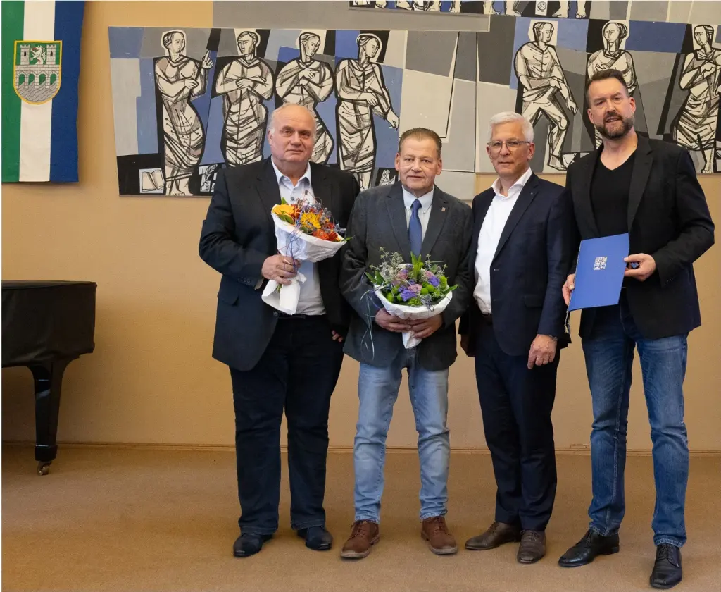Foto: Fünf Männer stehen in einem Saal eines Rathauses, vier halten einen Blumenstrauß in den Händen. Die Männer stehen mit Abstand zueinander.