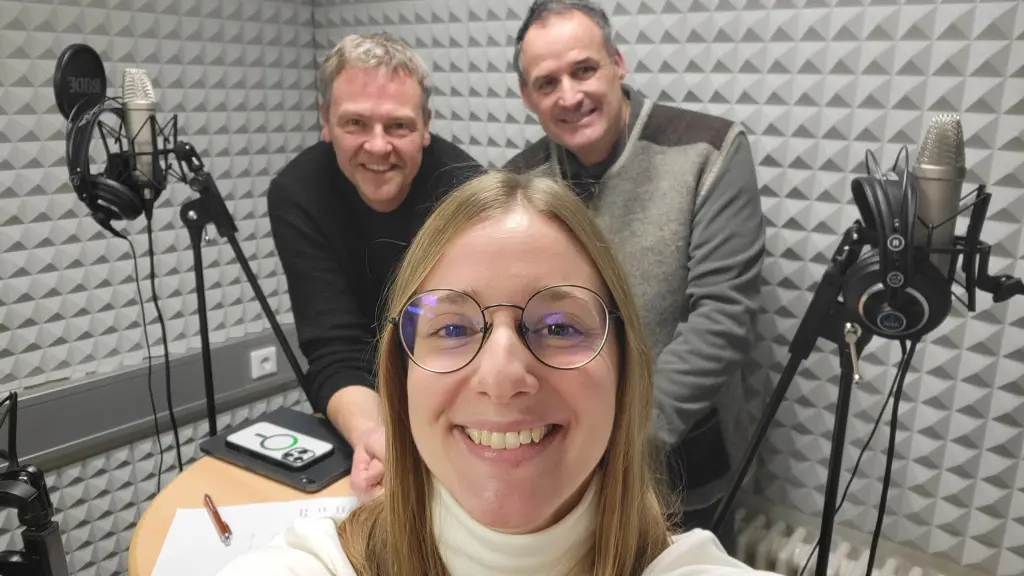 Drei Personen, die in einem Tonstudio stehen. Links und rechts im Bild sind Mikros und Kopfhörer zu erkennen.