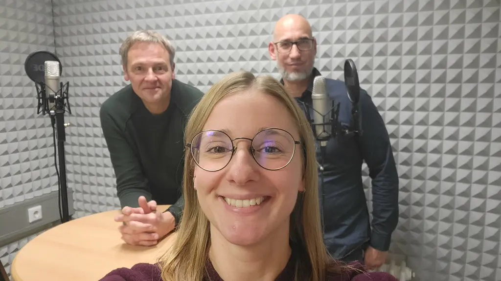 Drei Personen in einem Tonstudio, zu sehen sind außerdem Podcastmikrofone und Kopfhörer.