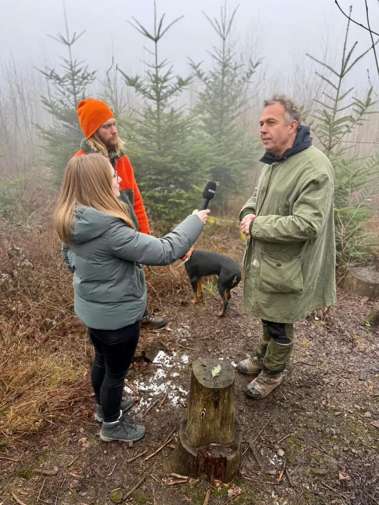 Im Vordergrund des Bildes ist eine Interviewsituation zwischen einem Mann und einer Frau im Wald zu sehen. Im Hintergrund steht ein weitere Mann mit seinem Hund.