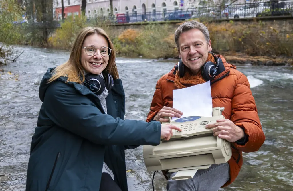 Foto: Die Podcast-Hosts Charlien Schmitt und Michael Kaub sitzen mit einem alten Faxgerät am Ufer des Flusses "Volme".