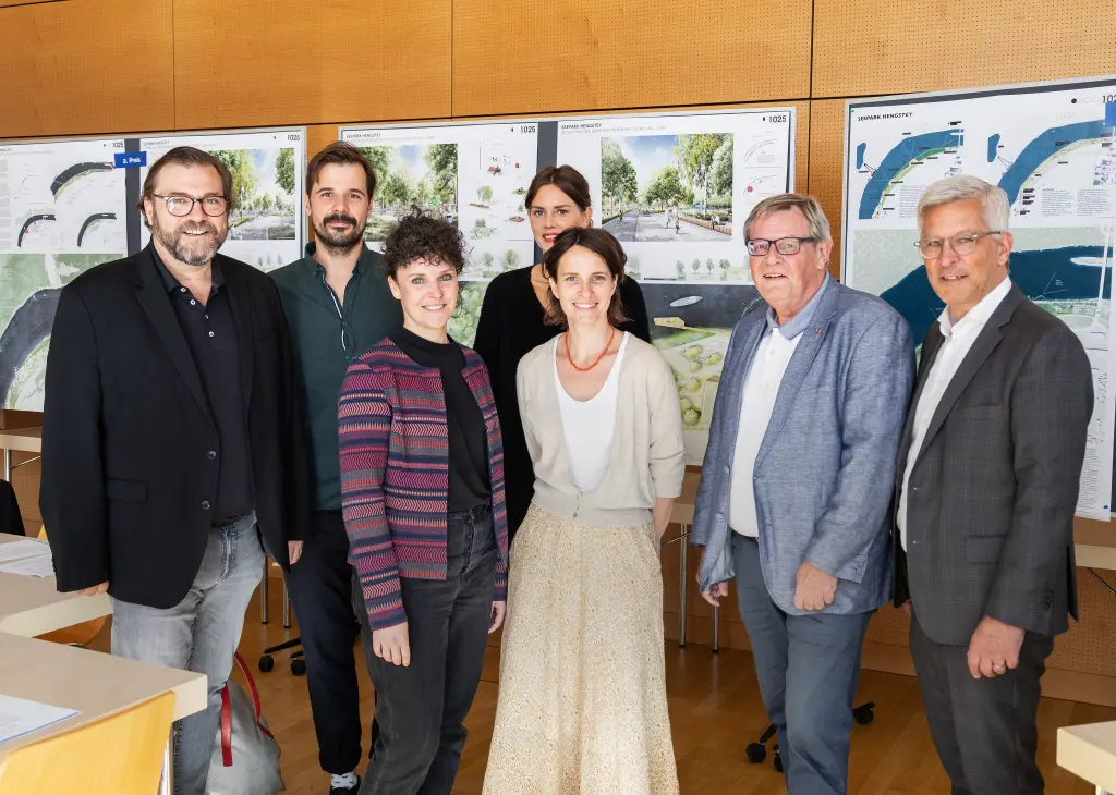 Foto: Die Preisträgerinnen und Preisträger des Architektenbüros zusammen mit Vertretern der Stadt Hagen stehen zusammen vor den Gewinnerentwürfen, die an die Wand hinter ihnen gepinnt sind.