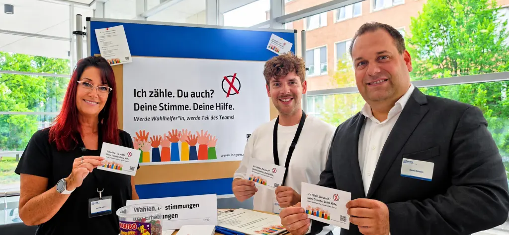 Foto: Informationsstand Wahlhelfer, Hintergrund:Der Text der Grafik ruft Wahlhelfer*innen auf, sich für die Tätigkeit bei der Stadt Hagen zu bewerben.