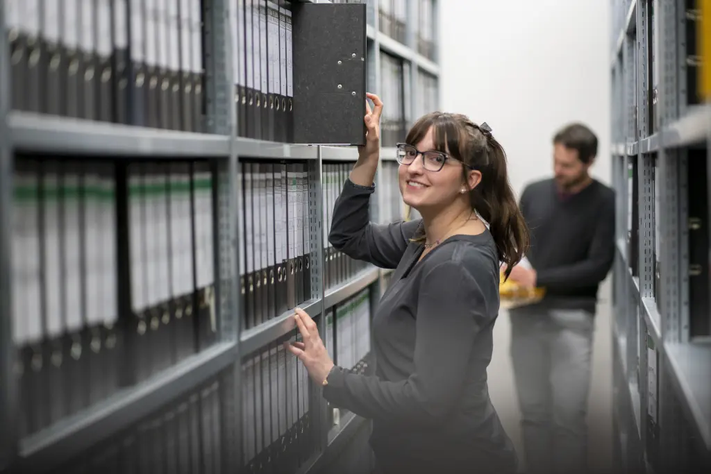 Eine freundliche Fachangestellte für Medien- und Informationsdienste im Bereich Archiv steht in einem gut organisierten Archivraum. Sie hat gerade einen Ordner aus einem Regal entnommen und schaut dabei freundlich in die Kamera. Ihre Haltung strahlt Professionalität und Engagement aus. Im Hintergrund ist ein männlicher Kollege zu sehen, der konzentriert in einem weiteren Ordner nach Informationen sucht. Der Raum ist mit Regalen voll archivierter Akten und Dokumente ausgestattet, was die strukturierte Arbeitsweise des Archivs unterstreicht. Die Szene vermittelt einen Eindruck von sorgfältiger Arbeit und Teamzusammenhalt, während die beiden Kolleg*innen effizient und fokussiert ihren Aufgaben nachgehen.