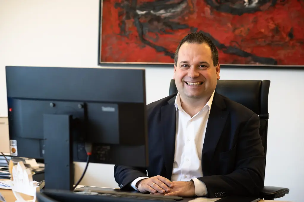 Foto: Dennis Rehbein sitzt an seinem Schreibtisch (ein Computerbildschirm sowie ein rotes Foto an der Wand sind zu sehen).