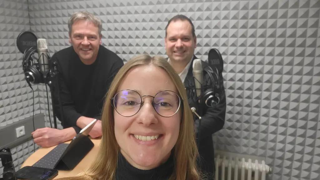 Drei Personen in einem Tonstudio. Zu sehen sind auch Mikros und Kopfhörer.