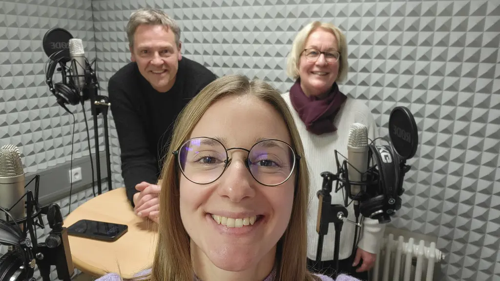 Zu sehen sind drei Personen in einem Tonstudio. Um Sie herum sind Mikrofone und Kopfhörer zu sehen.