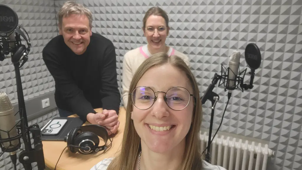 Drei Personen in einem Tonstudio. Außerdem zu sehen sind Mikrofone und Kopfhörer.