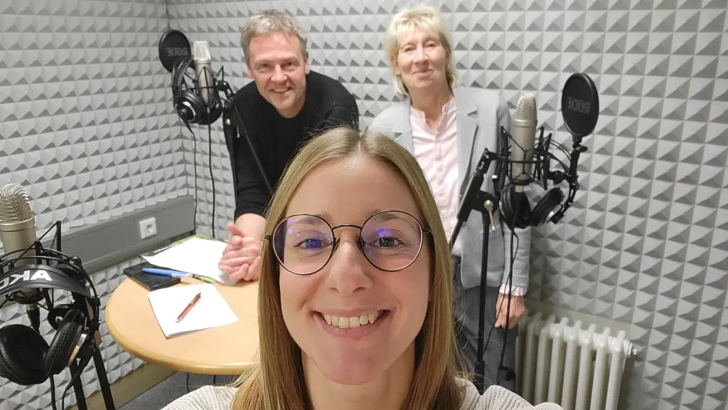 Drei Personen stehen in einem Tonstudio. Zu sehen sind Mikrofone und Mikrofonständer.