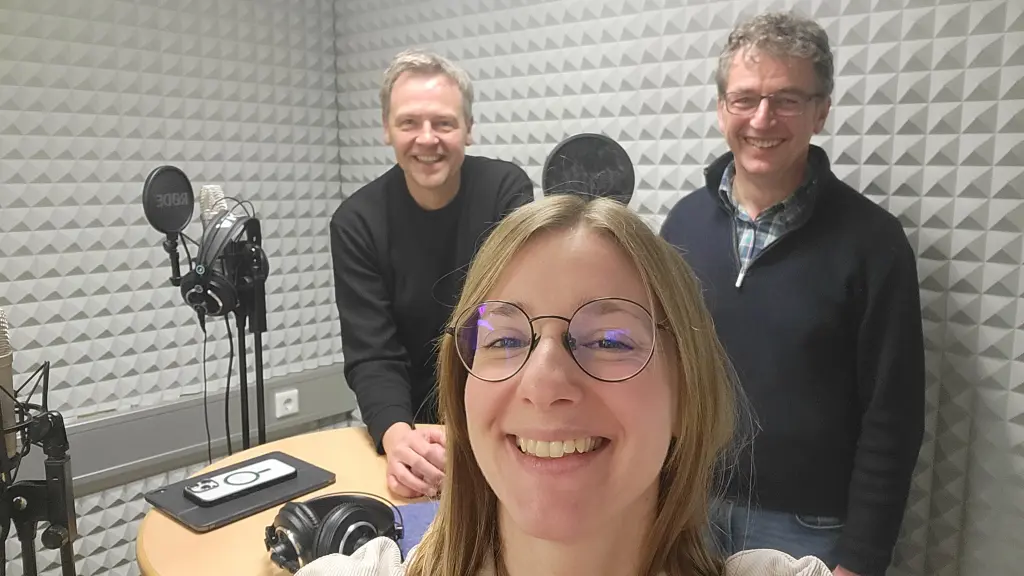 Drei Personen in einem Studio, zu sehen sind auch Mikrofone und Kopfhörer.