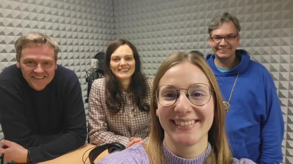 Vier Personen in einem Tonstudio. Im Hintergrund befinden sich ein Mikrofon und Kopfhörer.