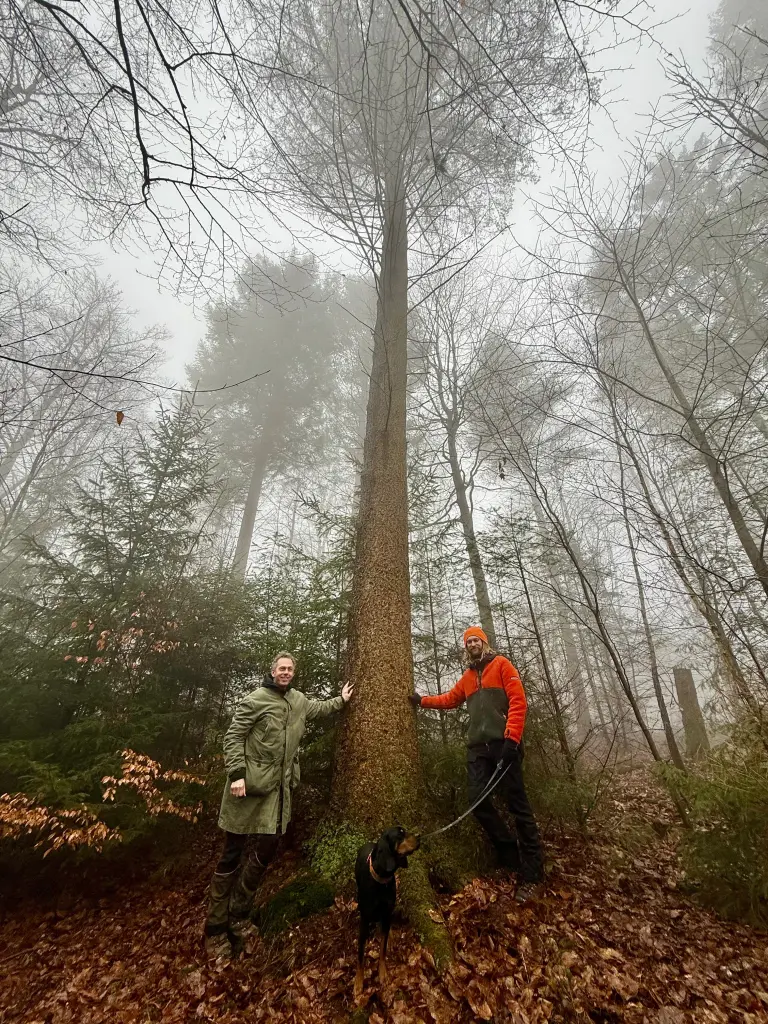 Zwei Männer und ein Hund stehen neben einem großen Baum.