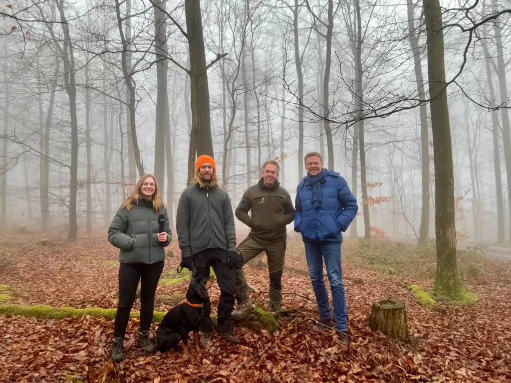 Vier Personen und ein Hund stehen gemeinsam im Wlad. Im Vordergrund befindet sich viel Laub. Im Hintergrund sind viele Baumstämme und Nebel zu sehen.