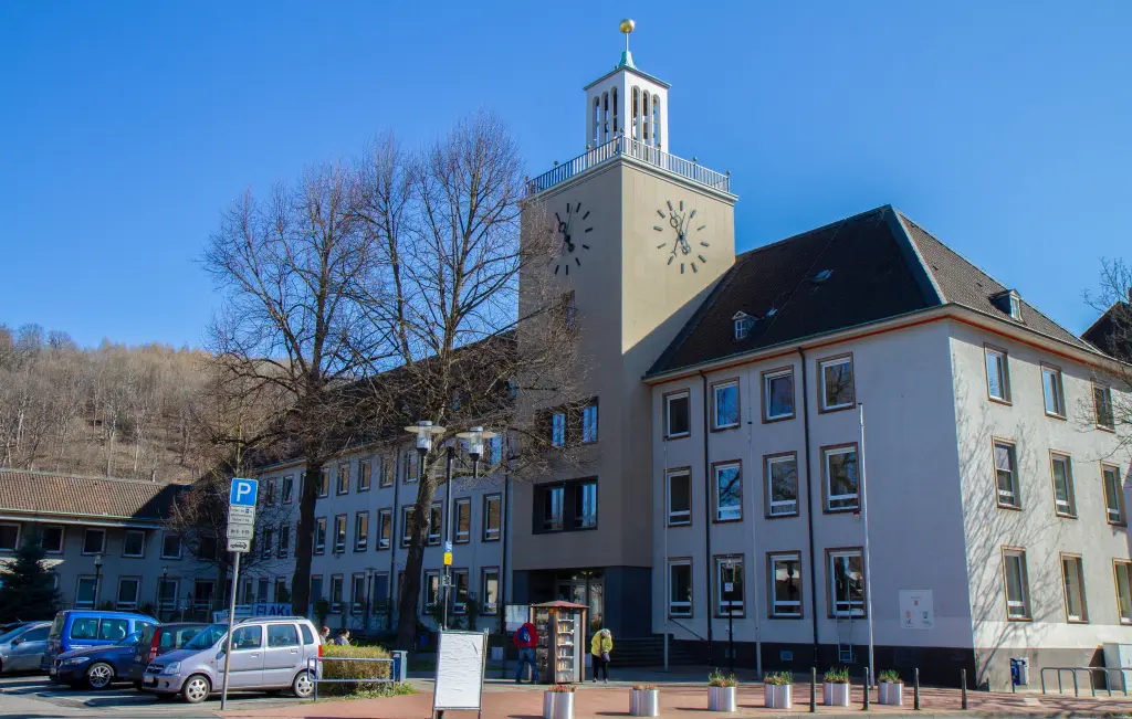 Foto: Zu sehen ist ein Rathaus mit Turm, davor befindet sich ein Parkplatz.