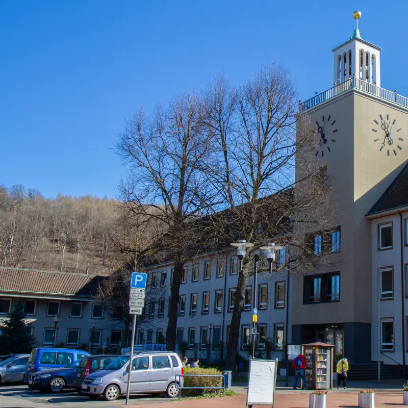 Foto: Zu sehen ist ein Rathaus mit Turm, davor befindet sich ein Parkplatz.