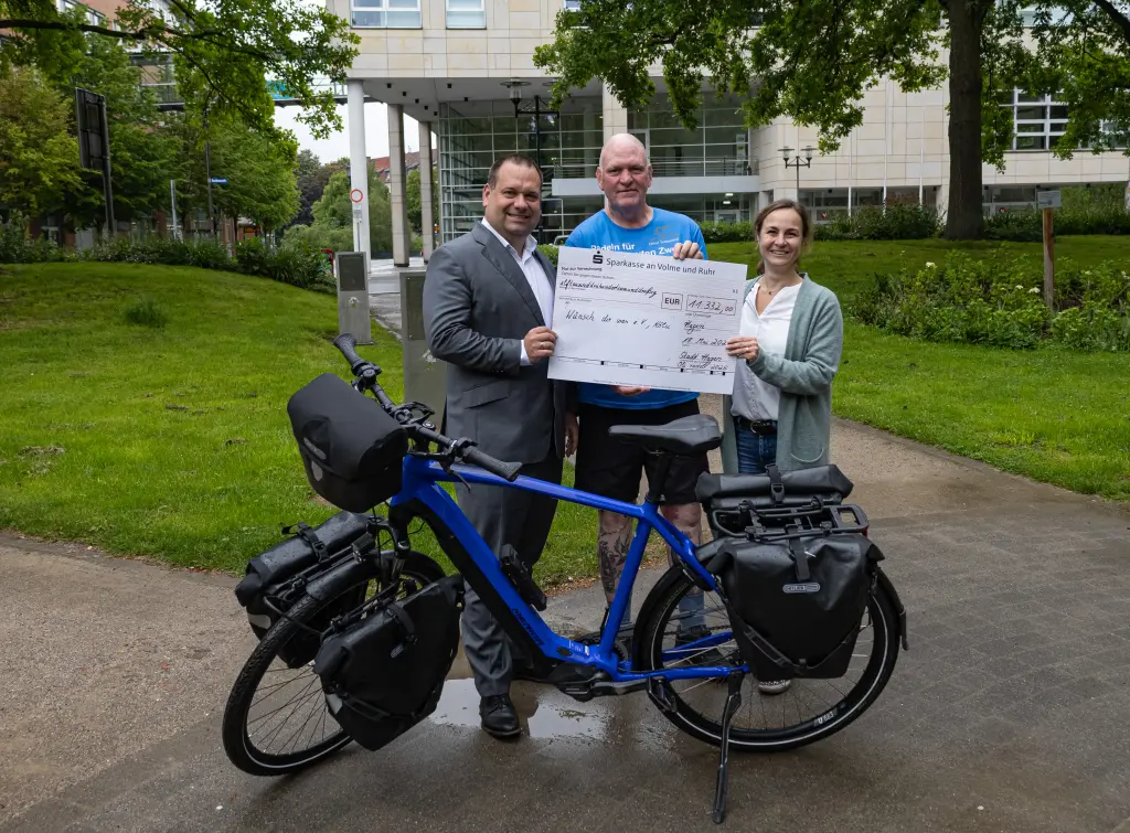 Foto: Oberb&uuml;rgermeister Dennis Rehbein, Oli Trelenberg und Nina Saure stehen zusammen vor einem blauen Fahrrad und halten einen Scheck in der Hand.