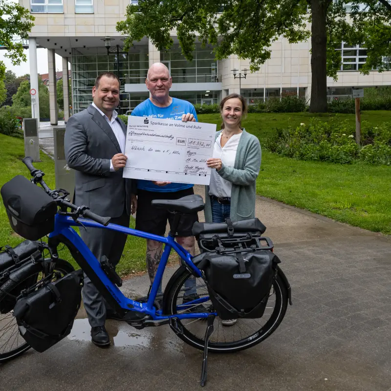 Foto: Oberb&uuml;rgermeister Dennis Rehbein, Oli Trelenberg und Nina Saure stehen zusammen vor einem blauen Fahrrad und halten einen Scheck in der Hand.