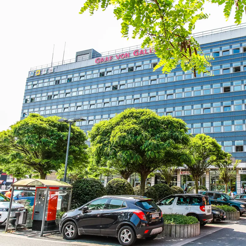 Foto: Ein großes Gebäude mit vielen Fenstern. Im Vordergrund befindet sich ein Parkplatz mit Autos und Bäumen.