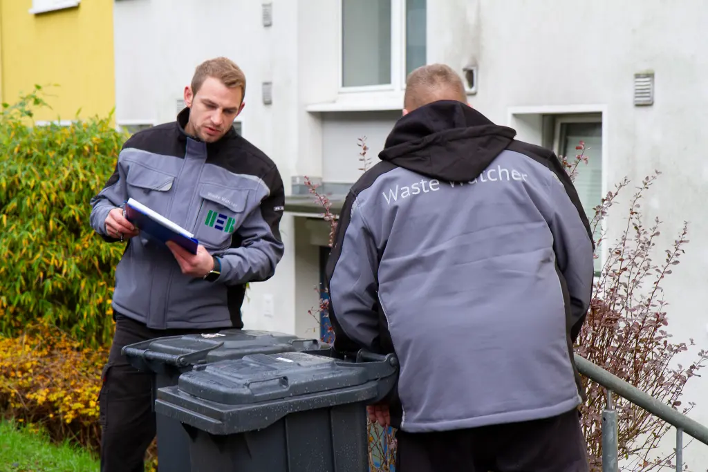 Foto: Zwei Männer in Dienstkleidung mit der Aufschrift "Waste Watcher" stehen vor einer Mülltonne. Im Hintergrund steht in Wohnhaus.