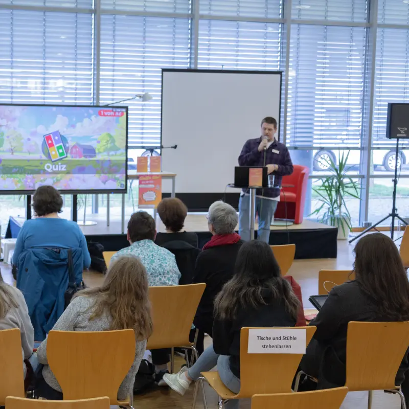 Foto: Mehrere Personen sitzen auf Stühlen und schauen auf den Fernseher, auf dem die Quizfragen angezeigt werden.