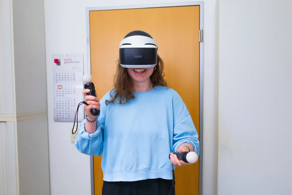 Foto: Eine Person hat eine VR-Brille auf und zwei Controller-Sticks in den Händen.
