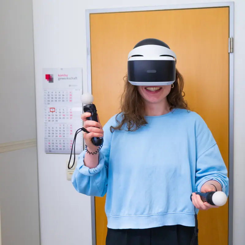 Foto: Eine Person hat eine VR-Brille auf und zwei Controller-Sticks in den Händen.